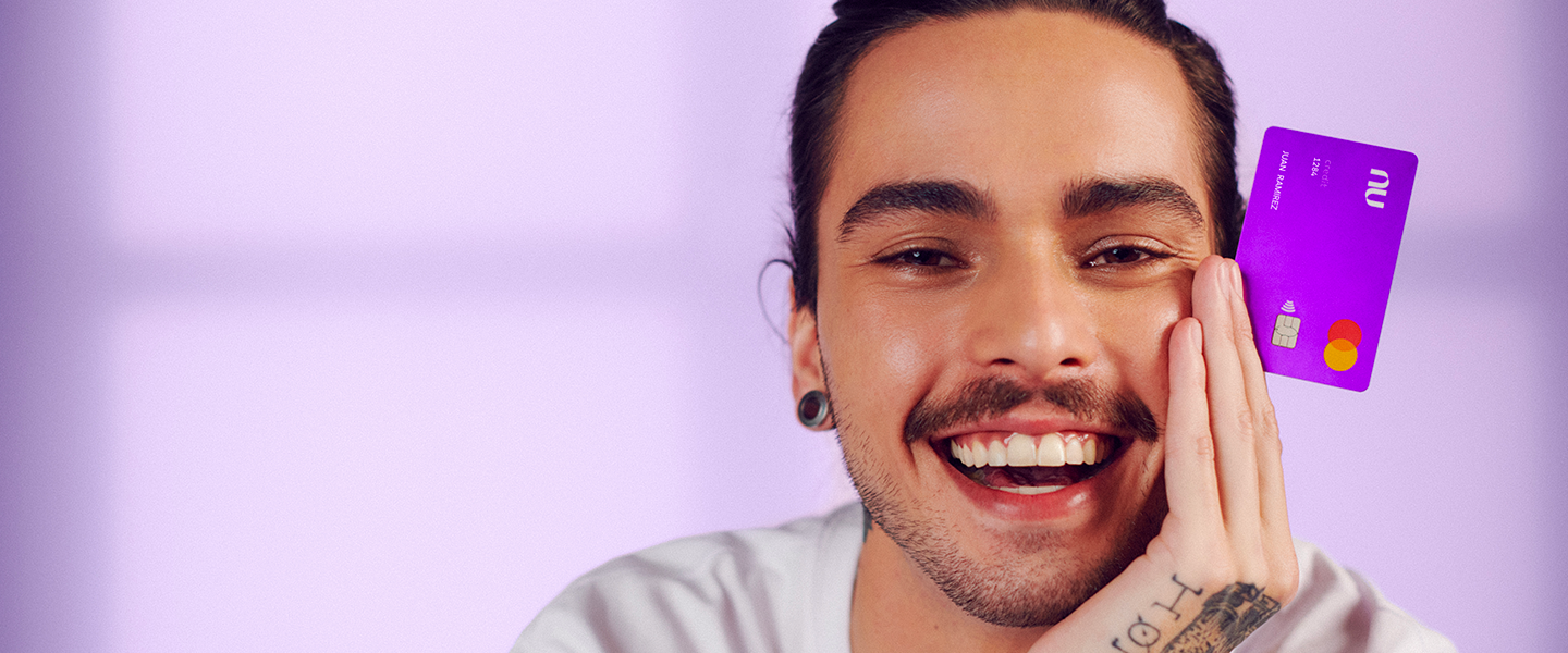 Young man with beard and tattooes on his hands smiles while holding a Nubank purple card