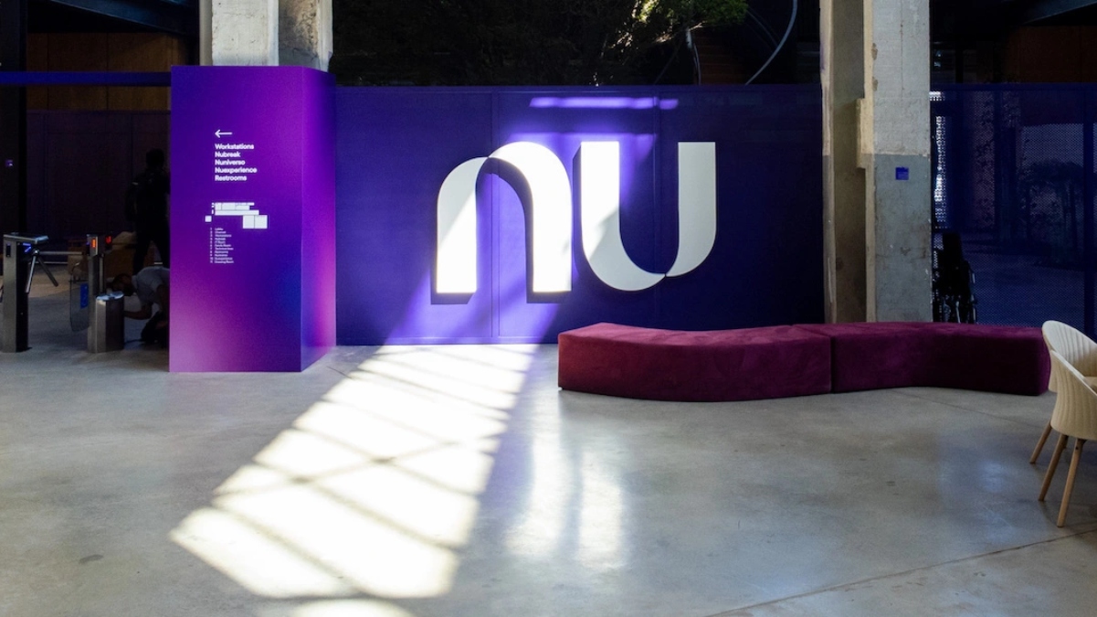Photo of the entrance to the new office at Nubank. You can see the very large white logo applied in front of a purple wall, a purple velvety fabric armchair and the turnstiles.